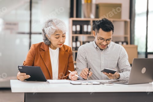Preview: An older mature woman talks to asian man. They have a laptop and papers. The man looks stressed.
