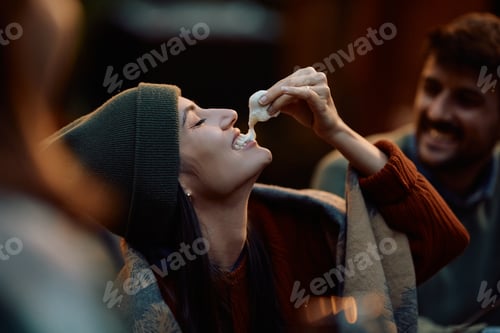 Preview: Happy woman tasting roasted marshmallows while camping with friends in autumn.