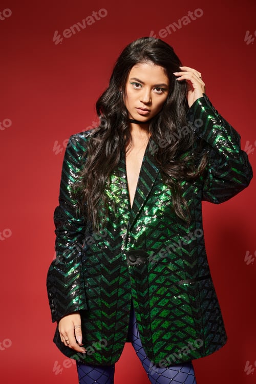 Preview: beautiful asian woman in trendy green jacket with sequins adjusting wavy hair on red background
