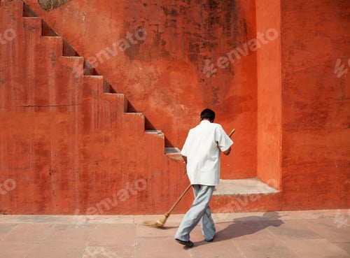 Preview: Rear view of man sweeping floor in front of red building with staircase.