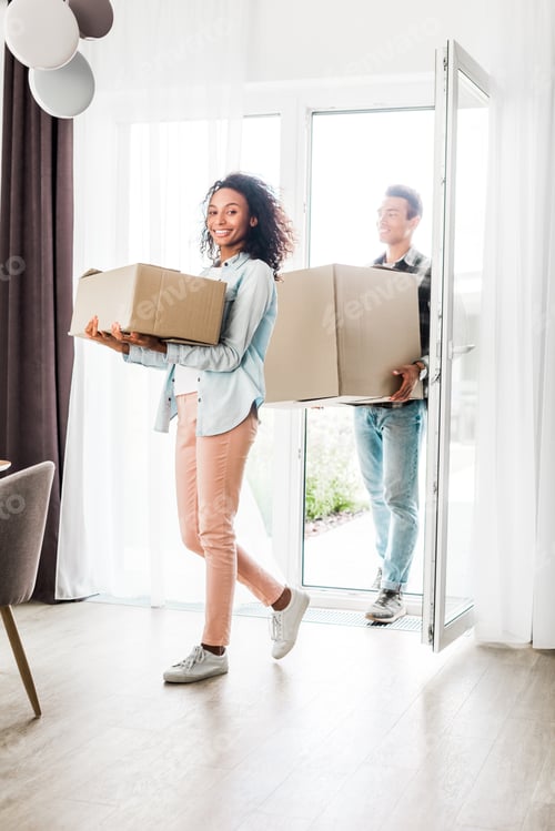 Preview: full length view of african american husband and wife smiling while walking into house, holding