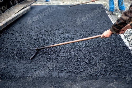 Preview: Worker leveling fresh asphalt on a road construction site, industrial building