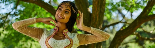Preview: cheerful indian woman in vibrant authentic attire posing and looking away in park, banner