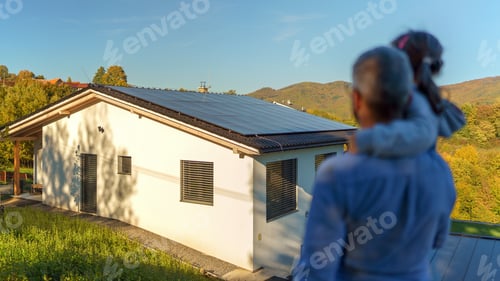 Preview: Rear view of dad holding her little girl in arms and looking at their house with installed solar