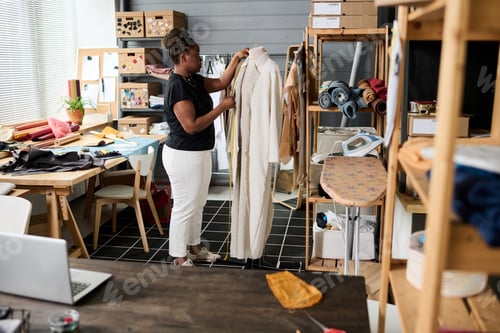Preview: Young African American female tailor standing in front of mannequin with coat on