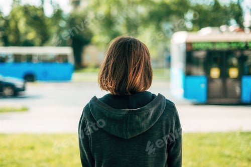 Preview: Rear view of adult brunette female standing on the street and looking at traffic and buses