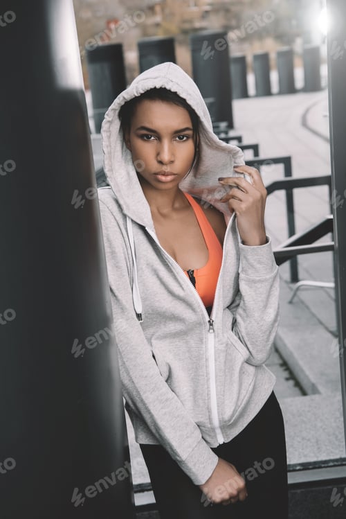 Preview: Attractive young sportswoman in hood posing on stadium and looking at camera