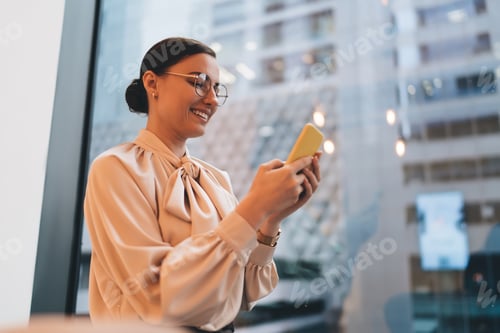Preview: Smiling female using smartphone standing near window