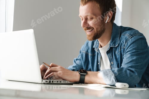 Preview: Photo of smiling man using wireless earphones while working with laptop