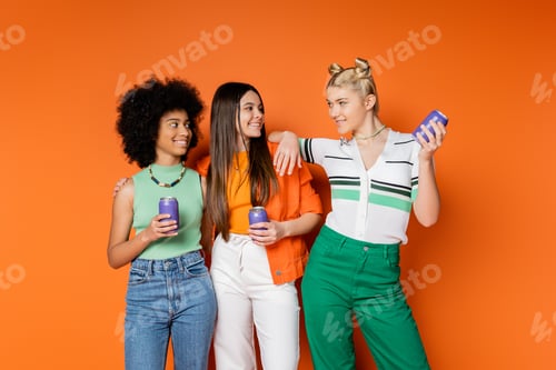 Preview: Positive blonde teenager in stylish outfit holding drink in tin can and looking at multiethnic