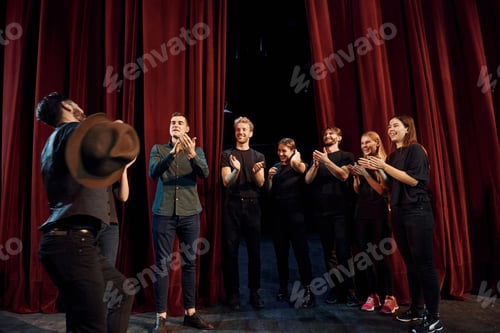 Preview: Standing against red curtains. Group of actors in dark colored clothes on rehearsal in the theater
