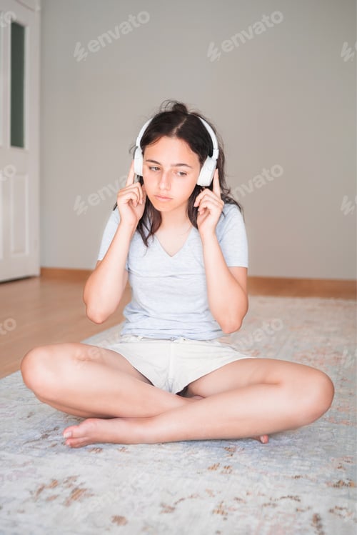 Preview: Pretty young girl listening to music with headphones while sitting on carpet at home