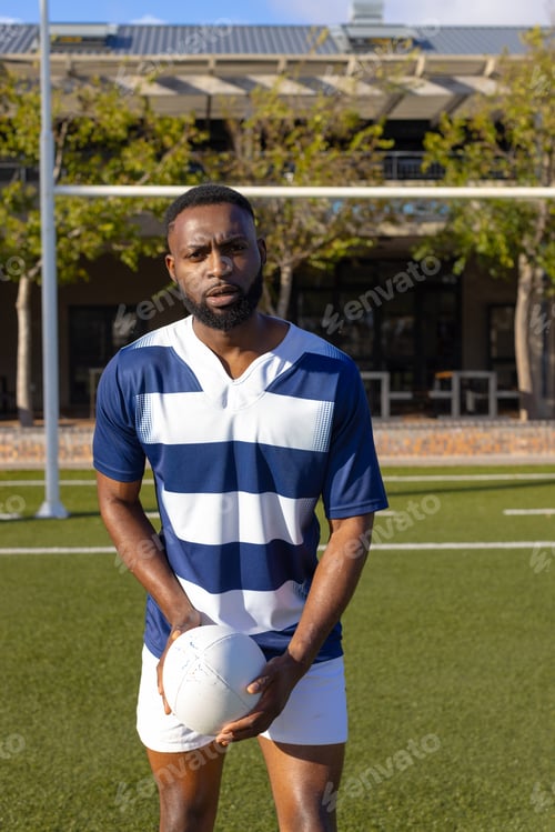 Preview: African American young male athlete holding rugby ball on field outdoors, looking serious