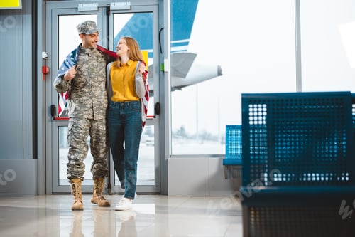 Preview: cheerful veteran in military uniform standing with girlfriend and holding american flag in airport