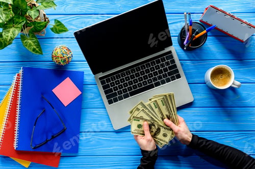 Preview: Business person counting dollars by laptop on blue wooden table with stationery