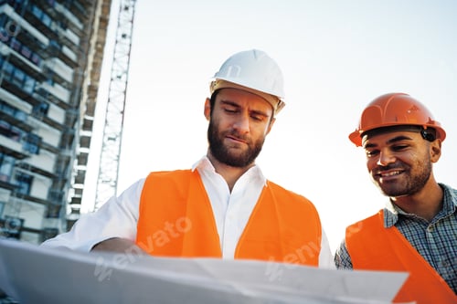 Preview: Two young male engineers in uniform and hardhats working at construction site