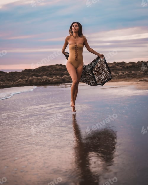 Preview: A brunette woman having fun with a swimsuit and a pareo walking along the beach in summer at sunset