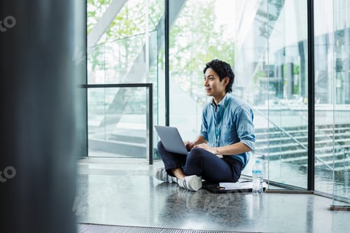 Preview: Businessman wearing blue shirt sitting on floor indoors, leaning against glass wall, working on