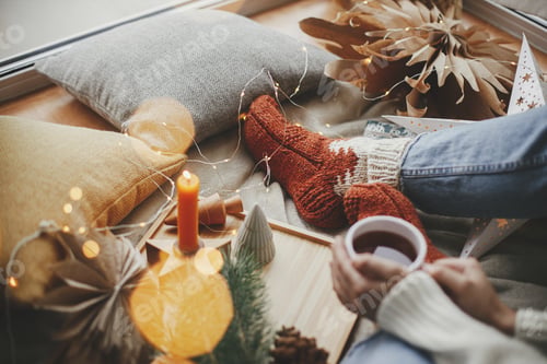 Preview: Cozy winter home. Woman hands with warm cup of tea on bed with lights, christmas star, pine, candle