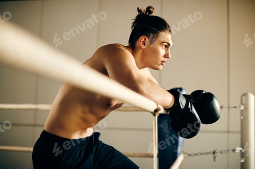 Preview: Young pensive boxer leaning on boxing ropes in a ring.