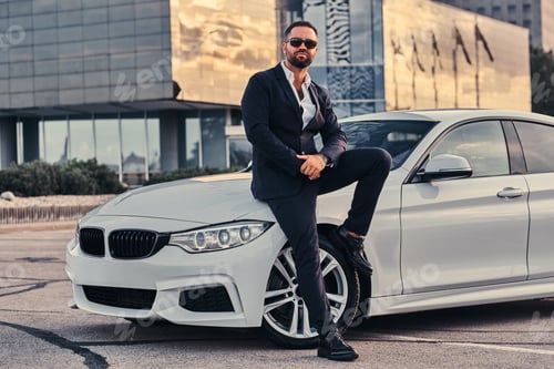 Preview: Bearded male in sunglasses dressed in a black suit sitting on luxury car against a skyscraper.