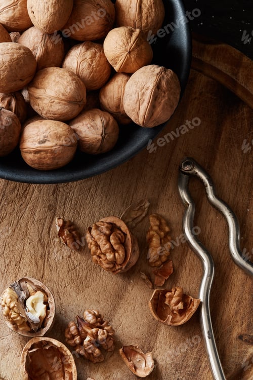 Preview: Cracked walnuts and kernels near the bowl with nuts and nutcracker on rustic wooden surface