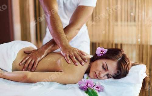 Preview: Purple flower lying down. Man does massage to the young woman in white towel indoors