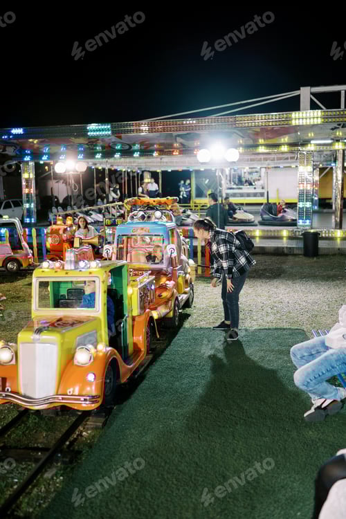 Visualização: Mamãe fica perto do vagão de trem infantil no parque de diversões e olha para a garotinha