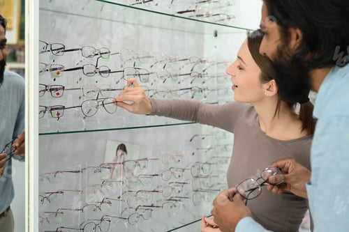 Preview: Beautiful female optician helping Indian man choosing eyeglasses frame in optical store.