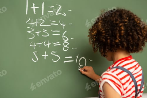 Preview: Schoolgirl solving mathematics calculations on green board in classroom at school