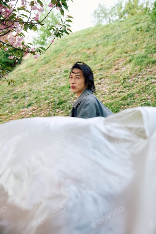 Preview: Young man in stylish grey outfit enjoys a serene moment in a park full of blossoms
