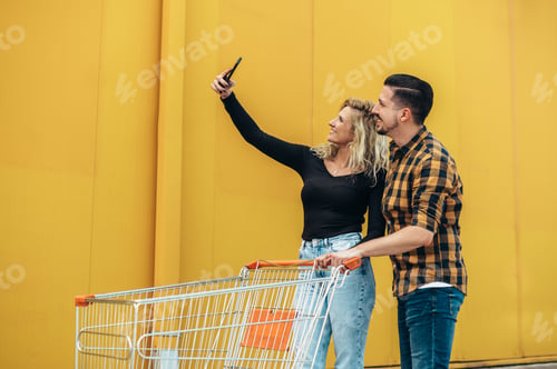 Preview: Couple using smartphone and taking a selfie while holding a supermarket shopping cart
