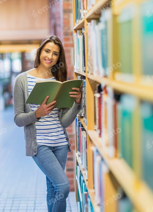 Preview: Woman in early twenties leaning in library aisle reading green hardcover book by wooden shelves