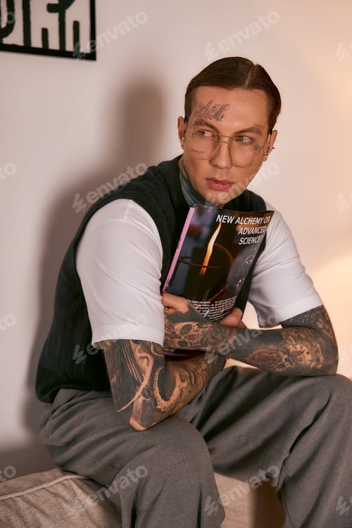 Preview: Handsome young man in stylish attire sits gracefully holding a magazine at home in the evening