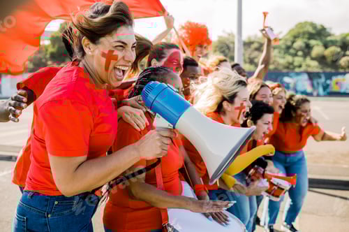 Preview: Female fan with painted face in foreground of group of fans cheering her team with megaphone.