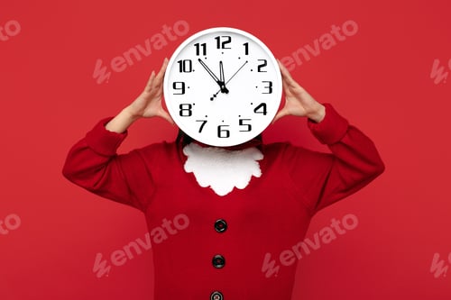 Preview: Woman dressed in Christmas santa sweater holding clock in front of face over red background.