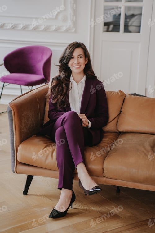 Preview: Vertical shoot of cheerful female manager sitting on leather couch dressed in violet suit at office.