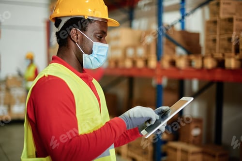 Preview: Caucasian adult warehouse worker checks delivery order on a tablet while wearing a safety mask