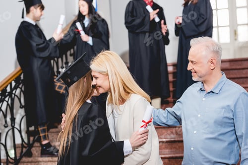 Preview: selective focus of girl in graduation cap hugging with mother near father