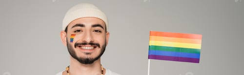 Preview: Cheerful homosexual man in hat looking at camera near lgbt flag isolated on grey, banner