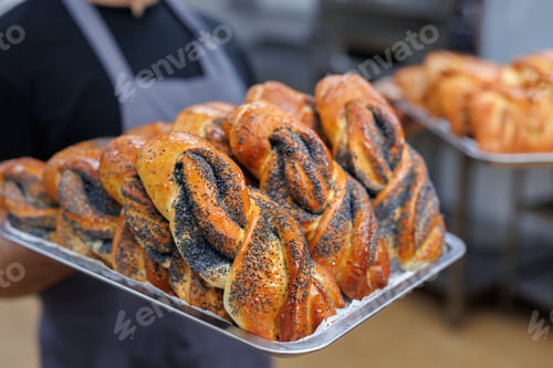 Preview: Baker carrying a tray of freshly baked poppy seed rolls in a kitchen.