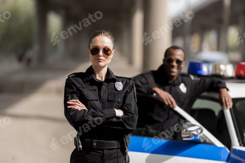 Preview: young serious policewoman standing with crossed arms while her partner standing near car and smiling