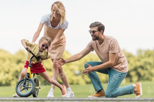 Preview: happy parents looking at cute little daughter riding bicycle in park