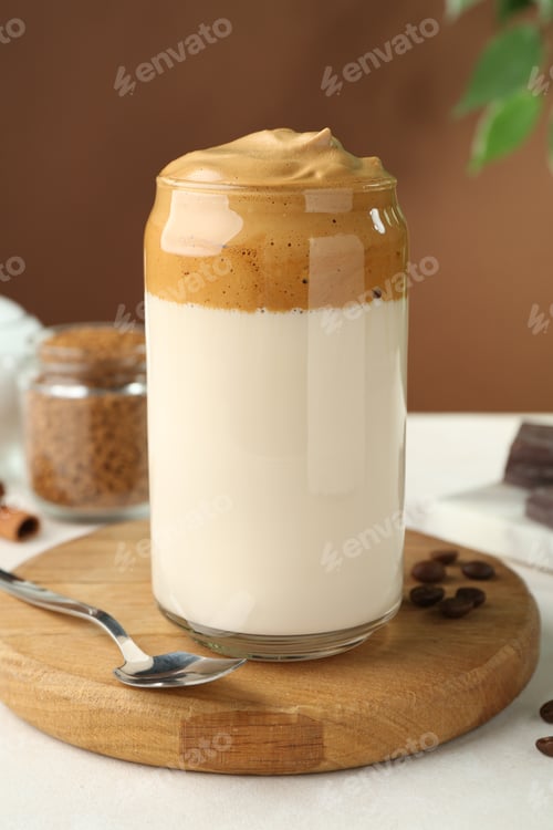 Preview: Delicious dalgona coffee in glass, beans and granules on white table, closeup