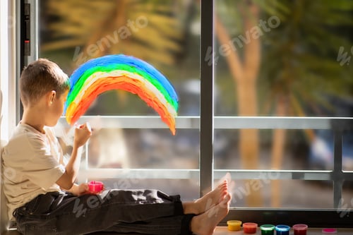 Preview: Busy caucasian little child sits on floor with paints, draws rainbow on panoramic window in