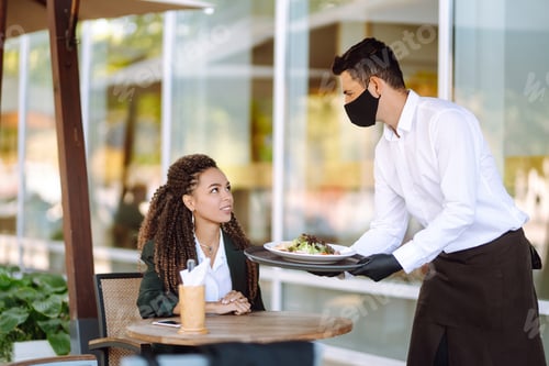Preview: Young waiter in protective face mask and gloves while bringing food to a customer in cafe. Covid-19.