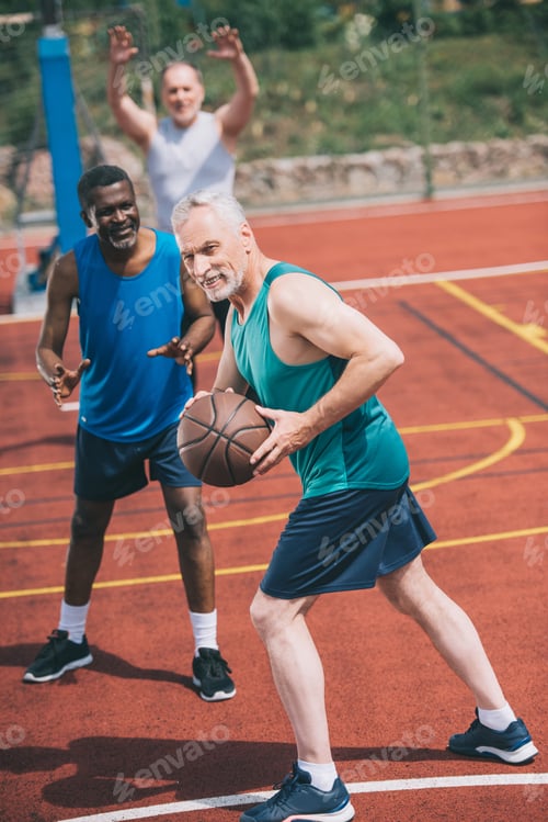 Preview: selective focus of old man with basketball ball in hands and multiracial friends behind on