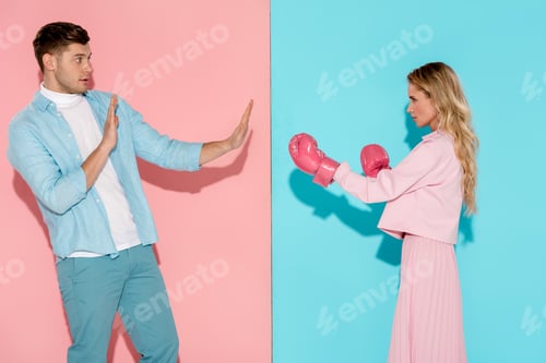 Preview: frightened man gesturing with hands near aggressive woman in pink boxing gloves on pink and blue