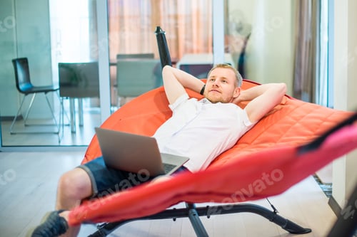 Preview: man with laptop holding hands behind head, relaxing and dreaming while lying on a hammock in office