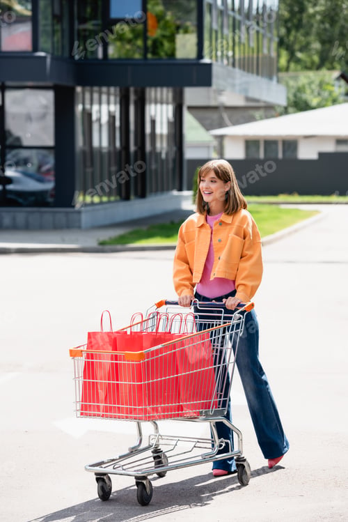 Preview: Woman in jeans and jacket looking away while walking near trolley with shopping bags on urban street